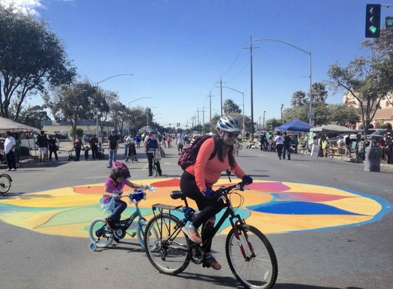 A woman and girl pedal around a mandala painted on East Alisal Street, Salinas, California. Behind them are people walking, biking, skating, scooting, and more on the car-free street enjoyed annually since 2013.