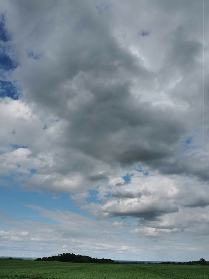 Cloud-filled sky with large, thick gray clouds dominating the upper part. A gap reveals patches of blue sky on the left. Below, a horizon with distant wind turbines peeks out between a stretch of green fields and a line of trees.