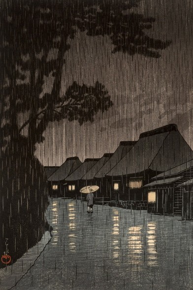 Evening rain falls on a rural street in Maekawa, in modern-day Kanagawa Prefecture. On our left, branches of tall shadowy trees reach out over the road; a row of traditional thatched-roof houses stands on our right, warm lights shining in their windows. The road running down the middle of the image is slick with rain, and steady rain continues to fall from a dim, overcast sky. A solitary pedestrian carries a bamboo umbrella over their head as they walk away from us.