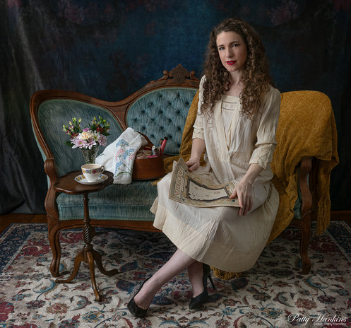 Woman in a vintage 1920s dress sitting on a settee looking at needlework magazine. Her sewing basket and needlework project are next to her on the settee