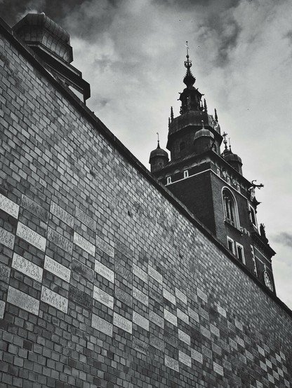 A low-angle, black and white shot of a brick wall on the left, adorned with numerous rectangular plaques inscribed with text. The wall recedes into the background, leading the eye towards a grand, ornate building with a prominent spire and multiple turrets, visible on the right side of the frame under a cloudy sky.