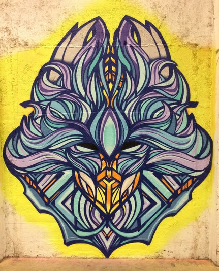 Symmetrical face representing a stylized oni demon face or mask done with spray paint on a wall. Flowing blue and purple organic shapes extend outwards from above the eyes and central third eye. Horns rise from the top of the head and a fiery orange glow can be seen emanating from within.