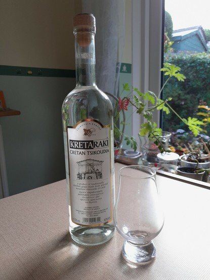 Unopened clear bottle of Cretan Tsikoudia (Raki) and an empty thistle shaped spirits glass on a kitchen table top.  In the background, a two-tone wall (yellow top, green lower) and pot plants in front of a glass door looking out onto a garden.