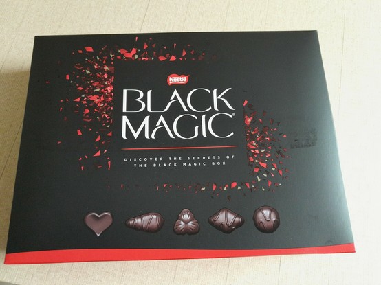 Unopened box of Black Magic chocolates: black background, white lettering, small red triangles form a design around the letters, and illustrations of 5 moulded chocolate designs.