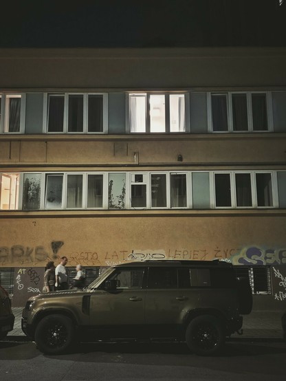 A night-time, eye-level, full outdoor shot shows a dark olive green Land Rover Defender parked on a street in front of a three-story building. The building has a mustard yellow facade, with two rows of rectangular windows. The top floor's windows are dark, except for one on the right that is brightly lit. The middle row of windows shows some reflections and wear. The ground floor has graffiti on its walls, and three people are visible near the car on the left side of the frame.
