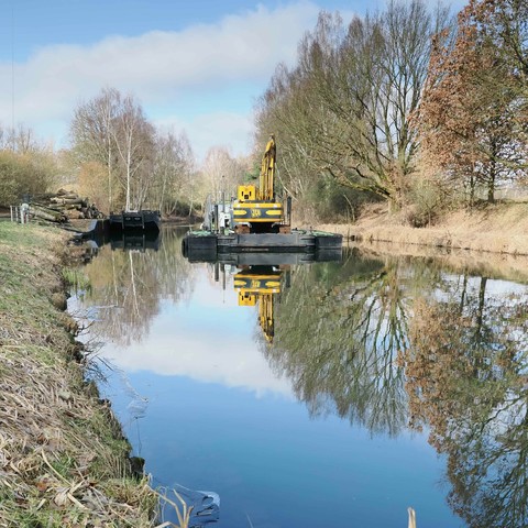 Excavator on a floating platform on a calm, narrow canal, surrounded by leafless and partially leafless trees reflecting in the water. A barge stacked with logs is near the excavator. The sky is partly cloudy, with blue and white reflections on the canal surface. Sparse grass lines the banks of the canal.