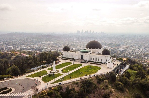 The image depicts the Griffith Observatory, a prominent landmark in Los Angeles, California. The observatory is characterized by its distinctive white architecture and two large domes, one of which is a prominent feature. The building is situated on a hill, offering a panoramic view of the city below. The foreground shows a well-maintained lawn with pathways and a few visitors scattered across the area. A circular road is visible, leading up to the observatory. In the background, the sprawling cityscape of Los Angeles is visible, with the downtown skyline faintly discernible in the distance. The sky is overcast, with a hazy atmosphere, suggesting a typical day in the city. The image captures the observatory's significance as a cultural and scientific landmark, with its location providing a vantage point over the urban landscape.

Provided by @altbot, generated privately and locally using Ovis2-8B