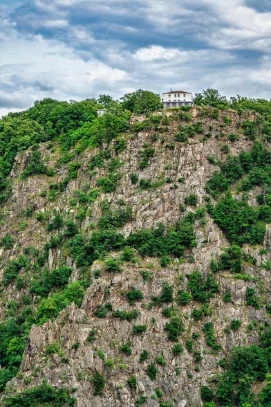 Felswand bewachsen mit Bäumen oben auf der Klippe steht ein weißes Haus und dahinter wolkenbehangener Himmel