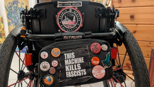 Un fauteuil roulant actif noir de dos avec un patch "M.V. LOUISE MICHEL SEARCH & RESCUE SOLIDARITY" sur le dossier. Une sacoche noire avec des badges, dont un grand "THIS MACHINE KILLS FASCISTS", est accrochée au fauteuil."