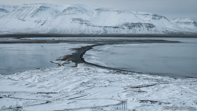 A colour photo which seems almost monochrome. On the horizon is a line of ice- and snow-clad mountains, their white flanks showing dark horizontal strata of black rock. The foreground is a land spit with a track winding sinuously down to the water's edge. The centre of the shot is a small bay with a stripe of black sand across it. On the right of it is the sea, on the left is a frozen lake. Waves are breaking sluggishly onto the right side of the black sand.