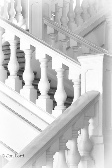 This is a black and white abstract photo of an all white staircase in portrait format. This staircase has three flights each about two to three metres long and at ninety degrees to each other. The steps of the first flight are out of view below with only the tops of the balustrades, handrail and newel post in view. The next flight turns ninety degrees to the left before turning ninety degrees to the right and disappearing from view at the top. Everything in the image is white. 
The location: Singapore (2012).
