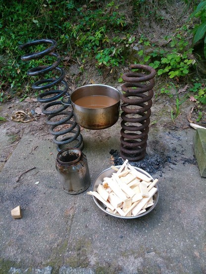 Ein kleiner Holzgaskocher steht vor zwei Auto- Spiralfedern, zwischen denen ein Kochtopf mit 1 L Kaffee hängt. Eine Schüssel mit 200 g Holz steht daneben.