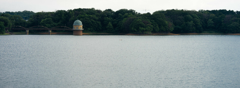 Calm body of water with a small, domed building on stilts near the shoreline, surrounded by dense, green trees. A bridge with multiple arches connects the building to the forested land. Overcast sky above, with the distant outline of hills in the background.