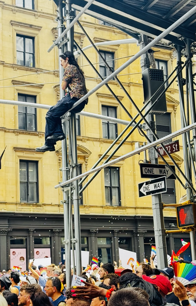 Color photo, in the foreground on the bottom are the tops of heads of people at the pride parade. On the corner is a great deal of metal scaffolding. A person has climbed up fairly high on the scaffolding and positioned himself nicely on the corner to get a full view of the parade. Behind him is a yellow building