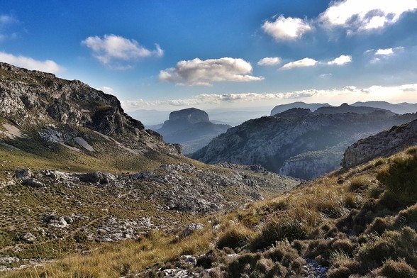 A wide-angle landscape shot of a vast mountainous terrain under a partly cloudy blue sky. The foreground features a steep, grassy and rocky slope leading into a valley. In the midground, rugged mountains stretch across the frame, with one prominent, flatter-topped mountain visible in the distance. Haze softens the far-off peaks, suggesting depth and a sense of scale. The overall mood is one of natural grandeur and tranquility.