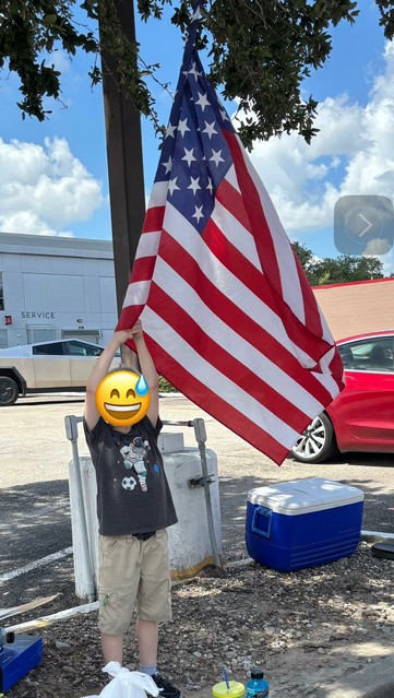 Young patriot holding an American flag, standing in front of a tree. In the near background is a blue cooler, and in the far backfound, on the left, a white, shaded building (the Tesla Service Center), and on the right, a sunlit brick buliding (Shipley's Do-Nuts).
