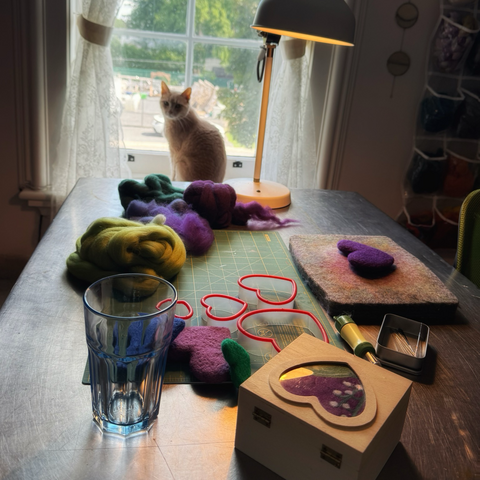 Stainless steel covered table in front of a window with dilute ginger cat sitting on window sill. On the table, there’s a lamp, green and purple wool roving, a green cutting mat, a wool felting pad, heart shaped cookie cutters, felting needles, felted hearts, an empty water glass, and a wooden box with a heart shape on top.