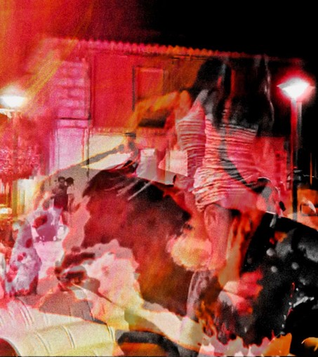 Color photograph in black and red tones, multiple exposures, motion blur. A woman rides an electric bull dummy, as if in a rodeo. The scene takes place at night in the village square of an old Spanish village, dimly lit. The superimposed exposure of several individual movements results in a blurred movement. The dominant blurred red and orange tones create an emotionally tense, heated atmosphere.