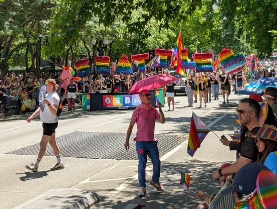 The GSBA group, with a colorful leading flag, marches in the distance, holding rainbow letters that spell out "Seattle❤️" Someone in the foreground is posed perfectly beneath the letters, wearing a pink shirt and holding a pink umbrella.