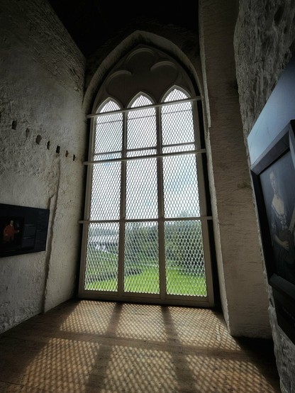 A low-angle interior shot within Tintern Abbey, Ireland, showing a tall, arched window with multiple panes and a diamond-shaped lattice pattern. The window casts intricate, patterned shadows on the wooden floor. Outside, a bright green field and trees are visible under a bright sky. To the left, a framed image hangs on a white brick wall, and to the right, another framed painting is partially visible, also on a white brick wall. The scene evokes a sense of historical tranquility and natural beauty.