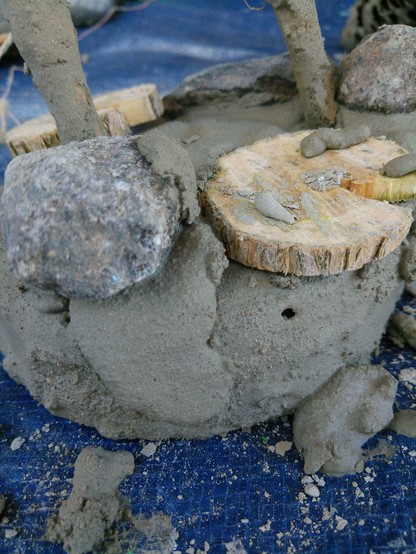 Eine in Beton gegossene Baumscheibe, mit Stein und einer kleinen Holzscheibe belegt, kleine Äste stecken darin.