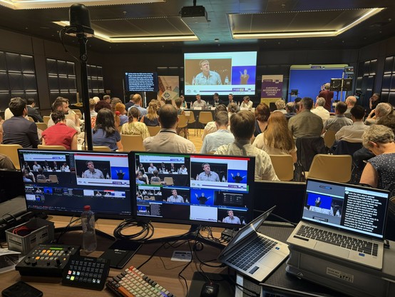 A view from behind of a conference room full of people. At the front of the room, several speakers are visible on stage, seated at the table with microphones, water bottles, and name tags in front of them. Behind the speakers, a large projection screen displays a close-up of Jakob speaking, along with a small window showing a sign language interpreter. Next to the screen is a banner for “European Accessibility Centre” and recognizable branding from the European Union. To the left side of the image, there is a large screen displaying real-time closed captions, 
In the foreground, a technical control station is set up with multiple computer monitors, keyboards, a switcher, and other audiovisual equipment spread across a long desk. The two central monitors display multiple live camera feeds of the event, including close-ups of speakers, wide shots of the panel and audience, and a view featuring the sign language interpreter. To the right, a laptop is running captioning software, showing the current captions that are being displayed both for in-room screens and remote viewers. Technical cables and small equipment are neatly arranged, with a water bottle.
