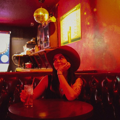 A woman in red lighting sits alone in a bar booth. She is wearing a cowboy hat and sleeveless black t-shirt. Her arms are covered with tattoos.