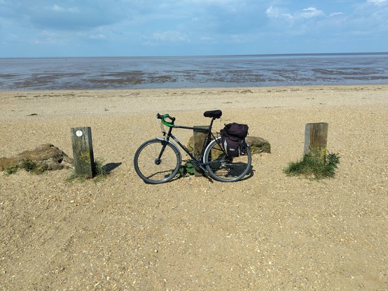 Una bicicleta negra con alforjas apoyada en un pequeño poste en la playa de Snettisham en Inglaterra. Se ve la arena y al fondo un terreno embarrado; el mar está retirado porque hay marea baja.