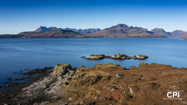 Dunscaith Castle ruins with a very blue sea (Loch Eishort) beyond then the Cuillin mountain range sitting below a blue sky. Isle of Skye, Scotland.