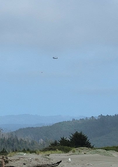 Helicopter with a water bucket beneath against a blue sky, with hazy mountains in the distance.
