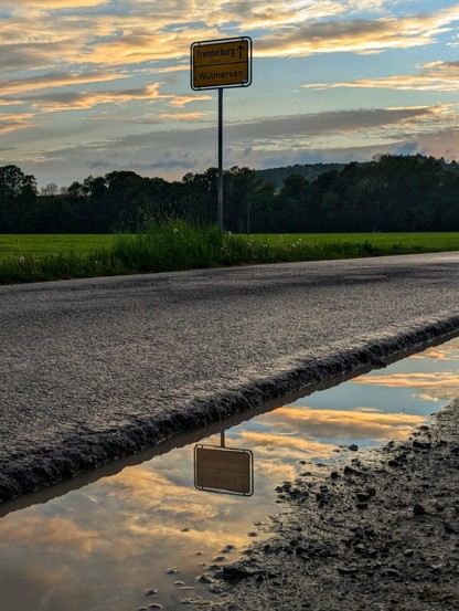 A low-angle shot captures a road stretching into the distance with a puddle reflecting the sky in the foreground. The sky is filled with dramatic, illuminated clouds at what appears to be either sunrise or sunset. A yellow signpost with black text indicates directions to "Trendelburg" and "Wülmersen". Green fields line the left side of the road, and a dark, dense forest stands on the horizon under the vibrant sky. The reflection of the sky and the signpost is visible in the muddy puddle next to the road.