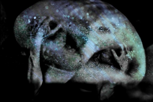 Color photograph in black and blue tones, landscape format, multiple exposure, self-portrait with small, round shards of glass from a shattered car window. The huddled, curled-up body of a person with their arms wrapped protectively around themselves. Double exposure with a very similar pose, mirror-inverted. Predominantly dark colors, covered with small, glittering greenish reflections. A single eye appears to gaze out at the viewer from within the body. The overall impression is gloomy but aesthetic, seeking security within, influenced by the outside world.