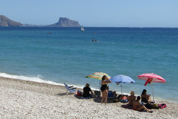 Am Strand liegen mehrere Personen mit 3 bunten Sonnenschirmen, dahinter das Mittelmeer und der Blickk auf den Felsen bei Calpe. Ein Segelschiff ist dazwischen auf dem Wasser.