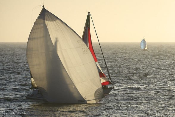 This is a colour sunset seascape photo of two sailing boats in landscape format. To the left of centre are two medium sized sailing boats with the closer boat healed over and overtaking the other but only with a short distance between the two of only few metres. The nearer boat's hull is almost all obscured by a very large white Genoa sail (Spinnaker) with only the stern visible. The Genoa is almost translucent with the setting sun causing silhouettes of the boats mast, rigging and sails visible through the sail. The sailing boat a short distance beyond is again mostly obscured by the white Genoa but the boat's bow and part of the bright red and white jib, visible. The sea surrounding our boats is dark blue and stretches upwards to fill three-quarters of the frame with a hazy horizon and a caramel coloured clear and cloudless sky above.
The location: Redondo Beach, California (2012).