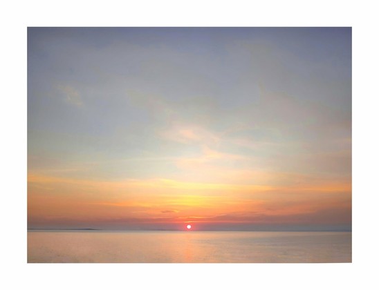 The photo is of a serene sunset over the calm ocean. The sun, a vibrant reddish-orange, sits low on the horizon and casts a warm glow across the sky. The sky is a blend of soft pastels - pale oranges, pinks, and blues - with thin, wispy clouds. The water is relatively still, reflecting the colors of the sunset.  There is a distant, dark shoreline which is barely visible on the horizon.
