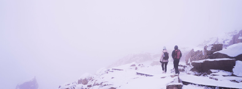 Two people dressed in winter clothing walk along a snow-covered, rocky path surrounded by dense fog. Jagged rocks border the pathway, and visibility is limited due to the misty atmosphere.