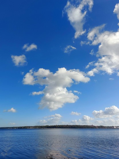 A sunny day by the sea, blue sky, calm blue waters, a flat coastline in the distance. Scattered high clouds like candy floss reflecting on the water.

Ein sonniger Tag am Meer, blauer Himmel, ruhige blaue See, in der Ferne eine flache Küste. Vereinzelte hohe Wolken wie Zuckerwatte, die sich auf dem Wasser spiegeln.
