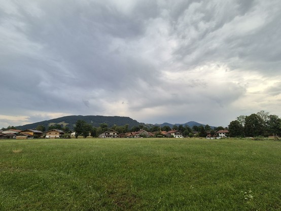 A serene and picturesque scene unfolds in this image, showcasing a large grassy field stretching out under a clear blue sky. In the distance, a cluster of houses can be seen nestled amongst the lush greenery. A solitary tree stands tall in the foreground, adding a touch of natural beauty to the landscape. The atmosphere is peaceful and inviting, with fluffy white clouds drifting lazily overhead. This idyllic rural setting is the epitome of tranquility, offering a glimpse into the harmony of nature and human habitation coexisting harmoniously.