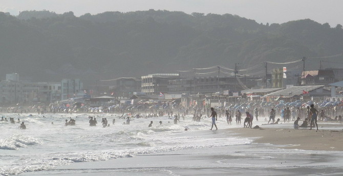 A long landscape photograph of a beach scene with white foamy waves. Distant crowds of people are in the sea and on the beach. One man is walking confidently into the sea. 

Further back are beachside businesses, fluttering flags, low-rise buildings and telegraph poles. Even further back are indistinct hills that loom over the scene. Spray from the waves adds colour perspective and the late afternoon sun is softened.