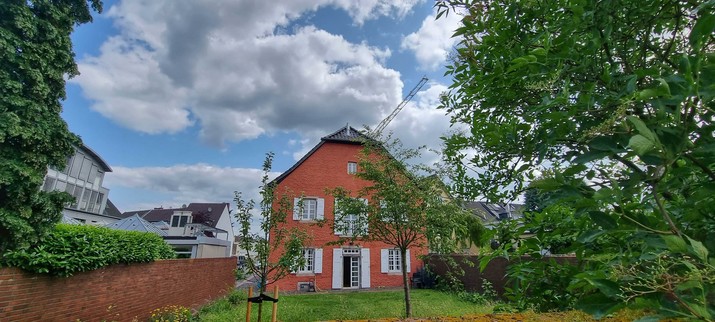 Blick in den frühsommerlichen Garten der Bücherei Süd in Mondorf. Blauer Himmel und ein rotes Backsteingebäude mit weißen Fensterläden. Das BücherWurmParadies wirkt sehr einladen.