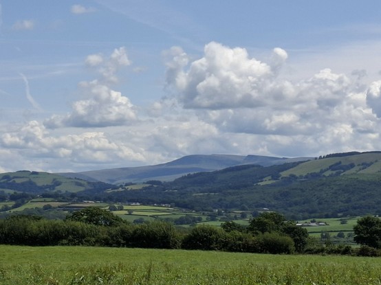 Looking across the valley of the river Twyi towards the Bannau Brecheiniog. The valley is a patchwork of foelds and trees. The sky is light blue with big fluffy clouds