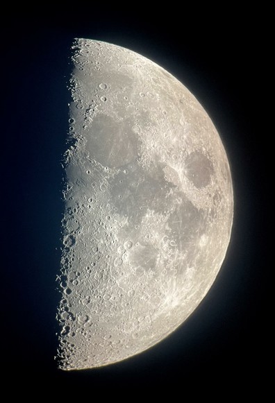 A first quarter moon against an almost black sky. Numerous craters are visible at the terminator, on the right, due to the low sun angle affording good shadow detail. Dark maria are prominent on most of the rest of the moon’s visible surface, against the lighter surrounding areas
