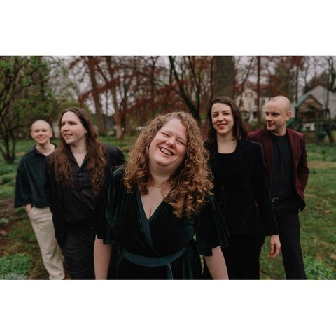 A group of five people dressed in black stand in a row in a grassy area. The woman in the middle, Brittany Ann Tranbaugh, has curly shoulder-length red hair and her head is thrown back in laughter.