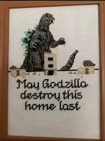 A photo of a needlepoint, cross-stitch in a wooden frame. Godzilla is portrayed walking up to a line of buildings. Under the buildings are the words “May Godzilla destroy this home last.”