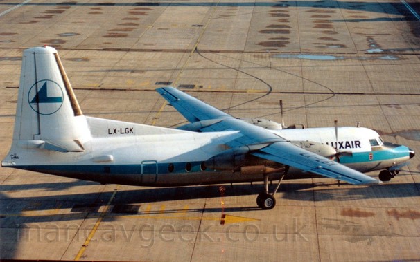 High side view of a high-winged, twin propellor-engined airliner facing to the right but being pushed back to the left by an out-of-frame tug attached to the nose-wheel by a long blue pole.
The plane is mostly white, with a grey belly, and a thick, light blue stripe running along the body.
There are black "LuxAir" titles on the upper forward fuselage, between the cockpit windows and the wing.
The tail is white, with the blue outline of a circle containing a rather angular, blue, stylised flying bird, and the black registration "LX-LGK" on a fillet at the front.
Grey concrete apron with occasional damp patches fill the rest of the frame.