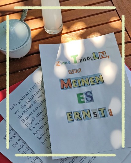 Auf der hölzernen Oberfläche eines Tischs, fotografiert von schräg oben, steht oben links eine weiße Zuckerdose aus Porzellan, unter dem Deckel ragt das Ende eines Löffels hervor, daneben steht eine schlanke weiße Blumenvase. Den größten Teil des Bilds nimmt ein DIN-A4-Blatt ein, auf dem mit ausgeschnittenen Buchstaben in verschiedenen Farben die Nachricht "Nicht trödeln, wir meinen es ernst!" aufgeklebt ist. Links unter dem Blatt liegen weitere Ausdrucke, deren Text man wegen des obersten Blatts nicht vollständig lesen kann. Das Bild ist von gelben Linien umrahmt.