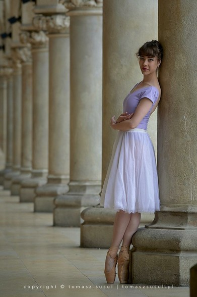 A ballet dancer stands en pointe, leaning gracefully against a large stone column in a corridor lined with classical pillars. She wears a light purple leotard and a white, knee-length tulle skirt. Her arms are gently crossed, and her posture is elegant and poised. The repeating columns and soft lighting create a serene, timeless atmosphere, emphasizing the dancer’s refined form and the architectural beauty of the setting.