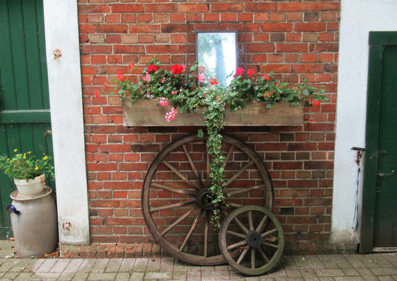 Ein Spiegel an einer roten Klinkerfassade, darunter ein Blumenkasten mit Geranien. Unterhalb zwei alte Kutschräder. Rechts und links Teile von grün gestrichenen Holztüren.