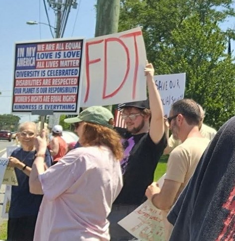 A group of men and women protesting by a roadway.  They are holding up signs for the passers-by to read.  One homemade sign reads, "FDT."  A printed sign in red , white and blue reads, "In My America:  We Are All Equal.  Love Is Love.  All Lives Matter.  Diversity Is Celebrated.  Disabilities Are Respected.  Our Planet Is Our Responsibility.  Women's Rights Are Equal Rights.  Kindness Is Everything."