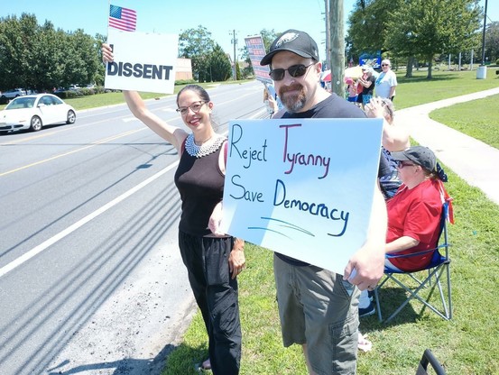 A woman protester, dressed as Ruth Bader Ginsberg, complete with black attire and her famous crocheted white collar, is standing by the roadside.  She is holding a small white sign with black letters that reads, "I dissent." Next to her is a man holding a sign that reads, "Reject Tyranny.  Save a democracy."